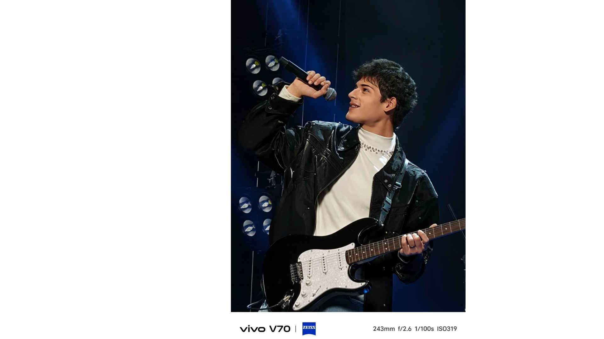 Male performer singing into a handheld mic while playing an electric guitar on stage with blue spotlight in the background